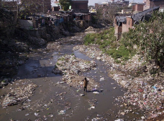 A photograph of a severely polluted river or stream running through what appears to be a slum area, with garbage piled along the muddy banks and a person standing in the middle of the debris-filled water.