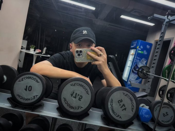 A young man wearing a New York Yankees cap takes a mirror selfie in a gym, posing behind a Technogym dumbbell rack displaying weights marked "10," "15," "2.5," and "3.5." In the background, a blue refrigerator and weightlifting equipment are visible, fitting the post's playful reference to his busy lifestyle in the crypto/Bybit community.