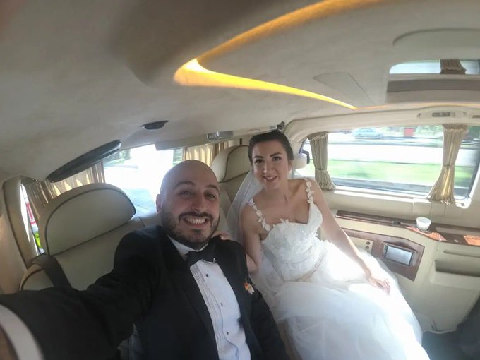 A smiling groom in a black tuxedo and bride in a white wedding dress sit together in the back of a luxury car with cream leather seats and ambient lighting.
