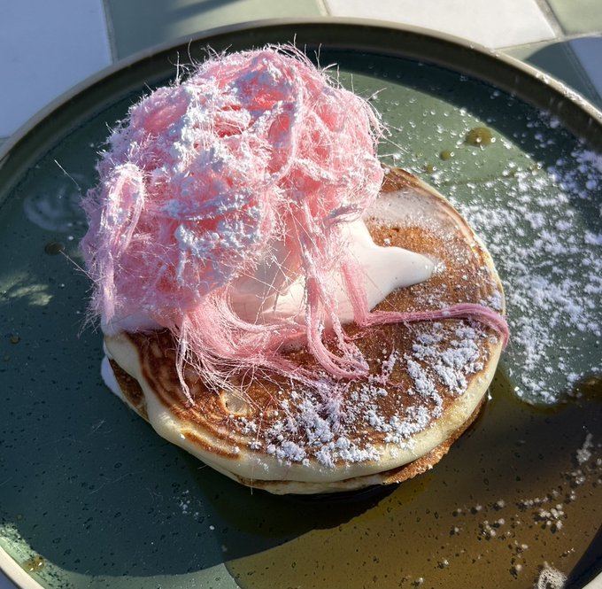 A close-up of a stack of pancakes topped with pink cotton candy and dusted with powdered sugar on a dark plate.