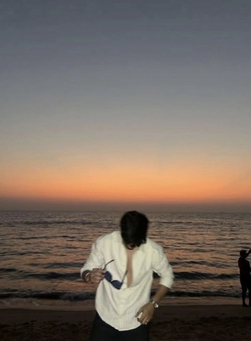 A young man in a white shirt holds sunglasses while standing on a beach at sunset, with another silhouetted figure visible in the distance against the ocean.