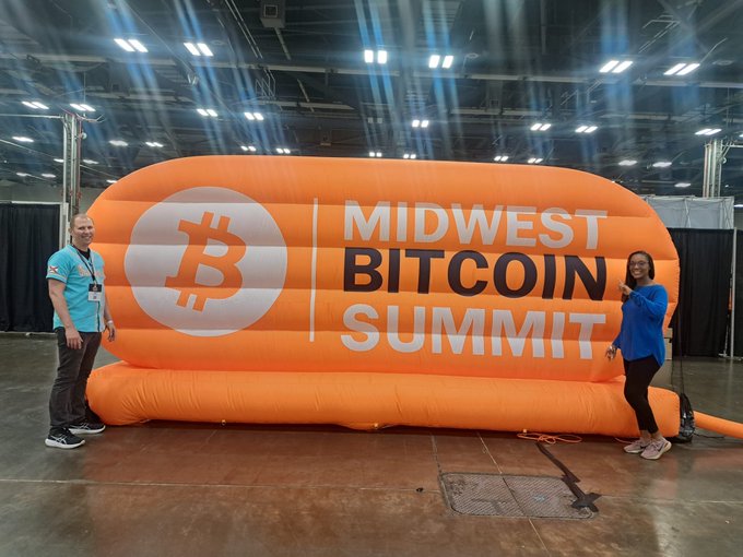 Two people pose on either side of a large orange inflatable Bitcoin logo banner that reads 'MIDWEST BITCOIN SUMMIT' at an indoor convention hall.