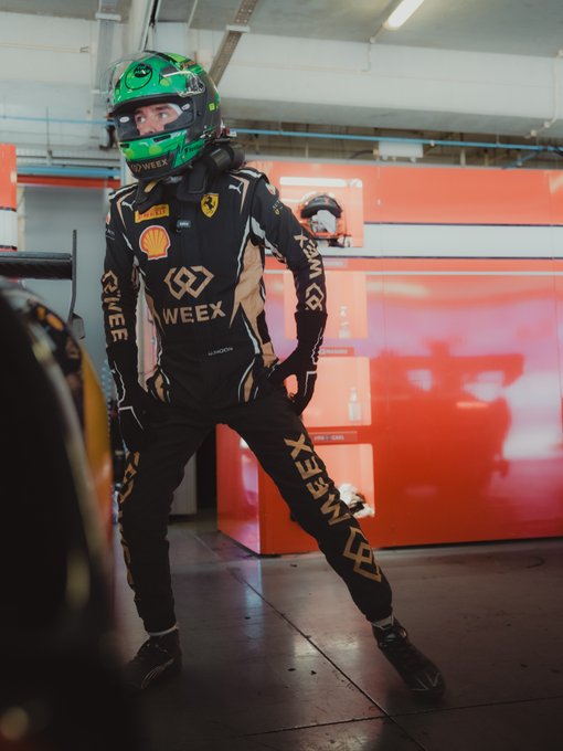 A racing driver wearing a green helmet and a black racing suit with WEEX and Shell logos stands in a pit garage area.