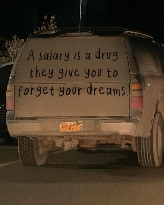 Nighttime photo of an SUV with hand-painted text on the rear window reading "A salary is a drug they give you to forget your dreams."