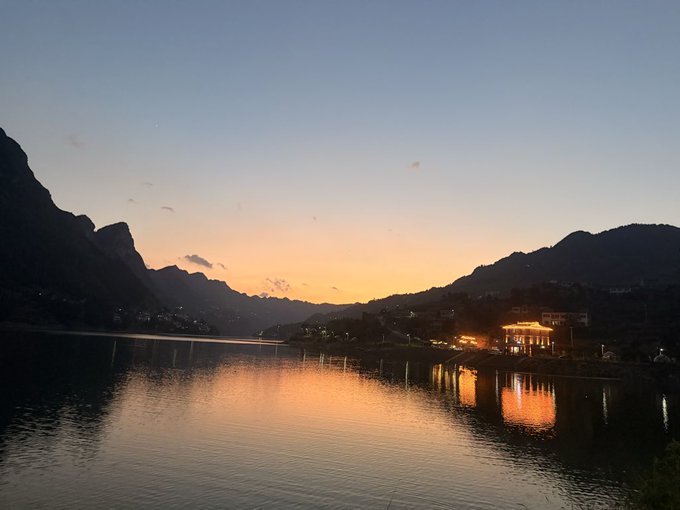 A serene landscape photograph of a lake at sunset or sunrise, with mountains silhouetted against an orange and blue sky and illuminated buildings reflecting on the calm water.