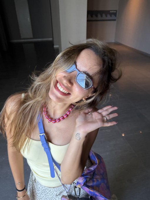 A smiling woman with blonde hair wears reflective sunglasses, a yellow tank top, and a chunky pink necklace, waving at the camera in what appears to be a modern indoor setting.