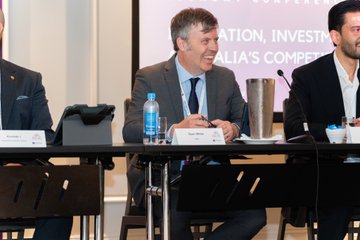 The image shows a group of three men sitting around a table at a trade conference. They are engaged in conversation during an event organized by the @XDC_Network. One man is located towards the left side, another one near the center, and the third one closer to the right side of the image. Various items are placed on the table such as water bottles, cups, and dining utensils like spoons and a fork. 

In addition to the physical objects, there is also a presentation displayed on the screen behind the men, indicating that this is likely part of an event taking place at the conference. The overall setting seems to be focused around digital trade, with discussions about investment strategies, market trends, and related topics being shared among the attendees.
