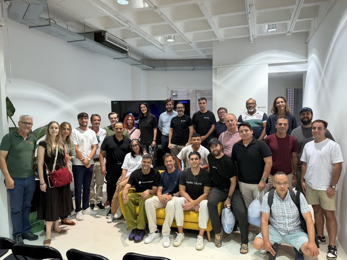 A large group photo of around 30 people posing together in a modern room with white walls and a yellow couch. Several people wear shirts with 'Hello Future' printed on them, including 'Hello, Tel Aviv' and 'Hello, Greece'.