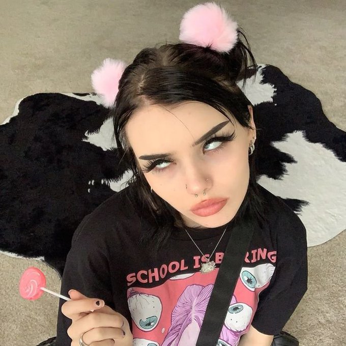 Portrait of a person with dark hair, heavy eyeliner, and pink fluffy ear accessories, wearing a black 'SCHOOL IS BORING' graphic t-shirt while holding a pink lollipop.