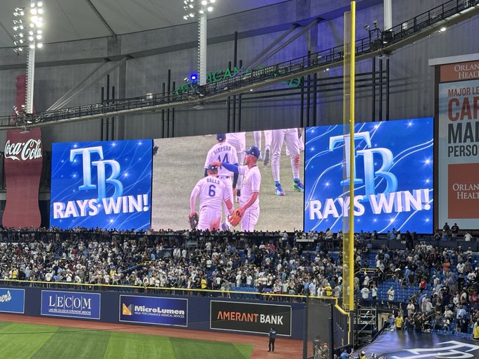 A view from the stands of a baseball stadium shows the Tampa Bay Rays winning, with large screens displaying "TB RAYS WIN!" alongside an image of player number 6, with sponsor ads for LECOM, MicroLumen, and Amerant Bank visible below.