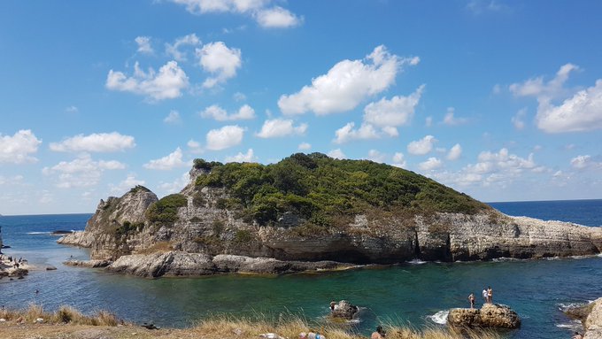 A serene coastal scene shows a lush, tree-covered rocky island surrounded by clear turquoise waters under a bright blue sky with scattered clouds, where a couple of people can be seen enjoying the calm sea. The image's tranquil, get-outside vibe pairs nicely with the post's "Happy Friday" energy, contrasting the natural beauty of the destination with the day's call to engage with @TheARCTERMINAL's AI tasks and privacy-focused mission.