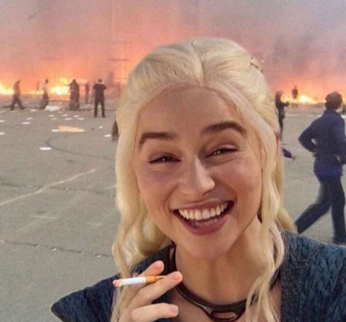 A blonde woman smiles while holding a cigarette in the foreground, with a chaotic scene of burning fires, smoke, and silhouetted figures in the background.