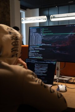 A person in a hoodie works at a desk with a laptop and monitor displaying code and charts.