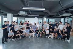 Group photo of a large team of people posing together in a modern office space with exposed ceiling pipes and large windows.