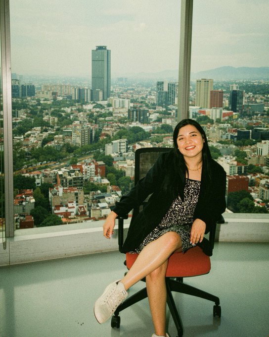 The image features a woman sitting in an office chair, smiling at the camera with her legs crossed. She appears to be wearing a black dress and white shoes. The background of the photo shows a cityscape with tall buildings, indicating that she is likely in an urban setting or within a large office building. The caption "In my corporate girl era! Bitso Offices #ETHMexico" suggests that this image might have been taken during her time working at a company named Ethos Offices.