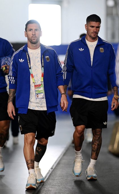 Two Argentine national team players, wearing official Adidas blue tracksuits with AFA crests and FIFA accreditation lanyards, walk through a stadium tunnel. The post references "Djed" (likely Rodrigo De Paul), connecting the image to the team's preparation as they prepare for a match.