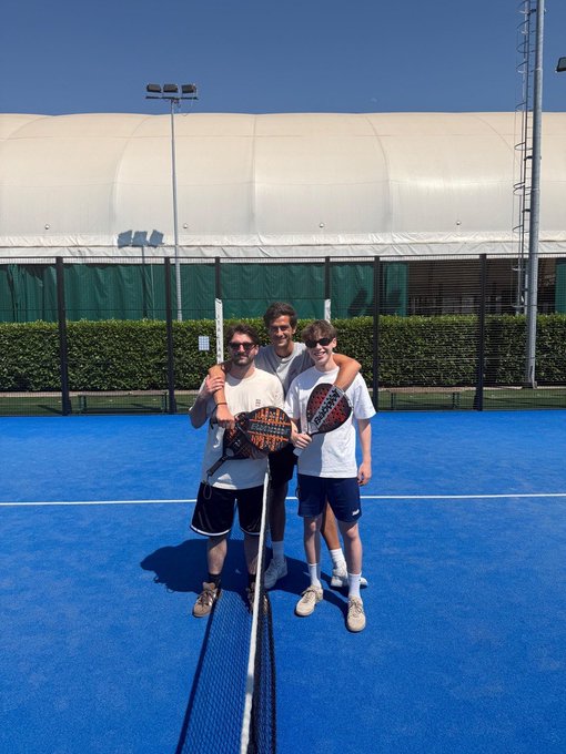 Three young men posing together on a blue padel court, with two of them holding padel rackets branded "adidas".