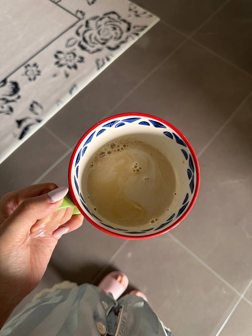 A hand holds a decorative blue and red mug of coffee above gray tile flooring, with a floral-patterned rug and pink slippers visible below. The image captures a quiet morning moment, reflecting the casual, behind-the-scenes vibe of the post about spontaneously setting up Bitcoin Asia meetups.