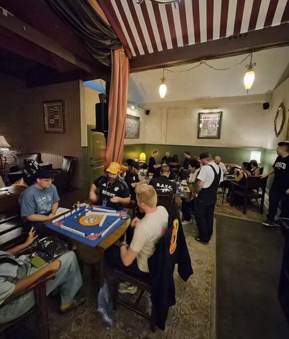 Indoor photo of a casual gathering where people are playing mahjong at a table in what appears to be a pub or lounge with striped awning ceilings.