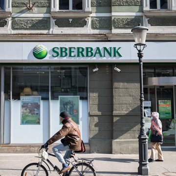 A man rides a bicycle past a Sberbank branch, while pedestrians walk along the sidewalk outside the Russian banking institution. The storefront displays the bank's green logo and name prominently above large windows featuring promotional posters, illustrating the backdrop for news that Russia's largest bank is expanding into cryptocurrency-backed lending.
