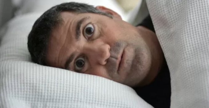 A close-up shows a man lying in bed with wide, startled eyes, his face partially covered by white bedding.