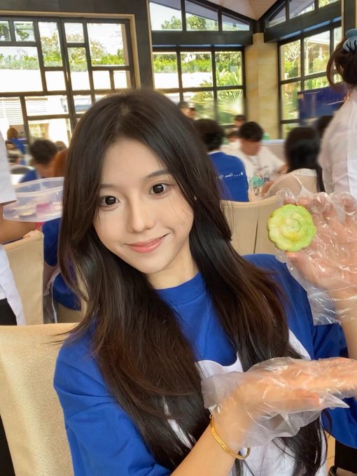 A young woman with long dark hair wearing a blue shirt smiles while holding up a small green and yellow pastry wearing a clear plastic glove, seated in a busy restaurant setting.