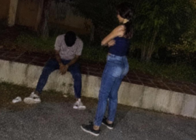 A nighttime photograph of a person in a blue tank top and jeans standing with arms crossed, facing another person sitting on a low wall in a white shirt and jeans.