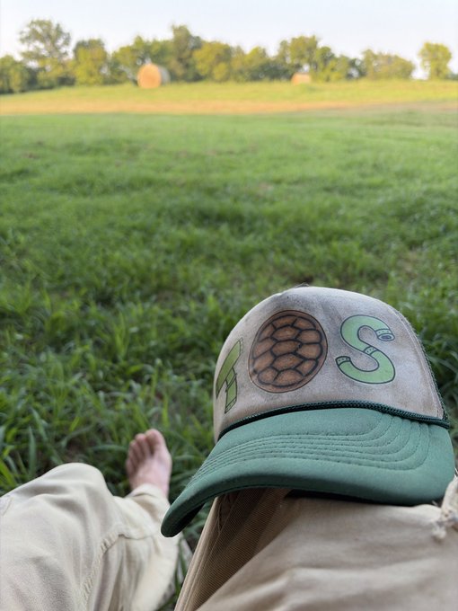 A first-person shot of someone relaxing barefoot on a grassy field with a "TOUCH GRASS" hat in view, capturing the literal act of "proof of grass" by touching the earth itself.