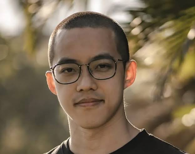 The image features a young man wearing glasses, standing outdoors with palm trees in the background. The man appears to be looking directly at the camera, giving off an air of confidence or curiosity. The caption provided seems unrelated to the main subject matter but is included for completeness and readability.