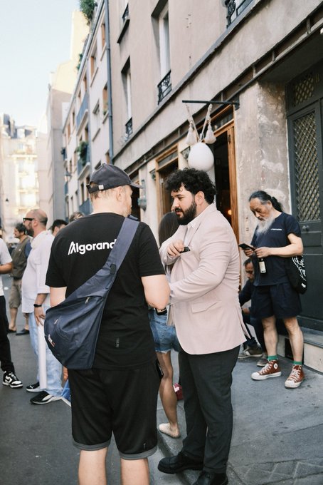 A group of people gather on a street, with one man wearing a black shirt displaying the "Algorand" logo and text on the back, while another man in a pink blazer and dark trousers stands facing him, and a third man checks his phone nearby. The scene reflects the post's mention of Algorand, with the branded apparel and business-attired individuals suggesting a professional or promotional event related to the Algorand blockchain platform.