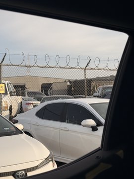 The image, taken from inside a vehicle, shows a parking lot secured by a chain-link fence topped with coiled razor wire, with several cars (including a white sedan and a Toyota) parked in front. The barbed wire and industrial setting align with the caption referencing the AST (AST SpaceMobile) site, suggesting the secured facility is being cleared out.