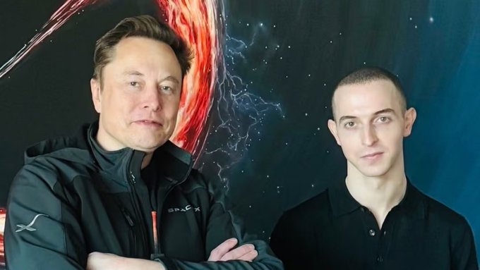 Two men posing in front of a space-themed backdrop featuring a fiery planet; the man on the left wears a SpaceX jacket, while the man on the right wears a black shirt.