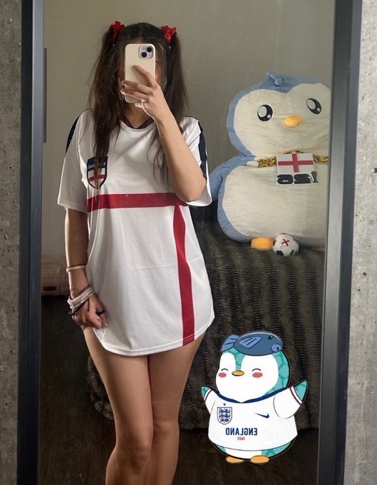 A person wearing a white and red England soccer jersey takes a mirror selfie with a phone, standing next to a large plush penguin toy with an England flag, while a penguin sticker labeled 'ENGLAND' appears at the bottom.