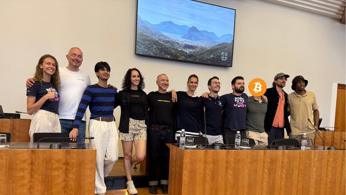 Group photo of eleven people posing in what appears to be a conference room, with a Bitcoin logo overlaid on the image and a screen displaying a mountain landscape behind them.