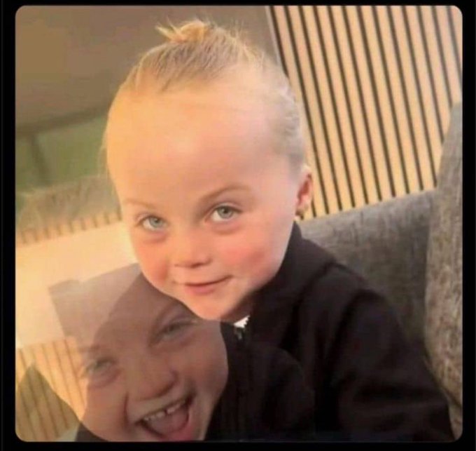 A vintage-style photo showing a young blonde child with hair pulled up, with an adult's smiling face visible beneath the child, against a background of vertical wooden slats.