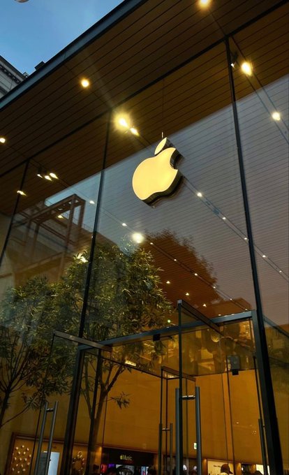 The illuminated Apple logo is mounted on the glass facade of an Apple Store at dusk, with interior lights and trees visible through the windows.