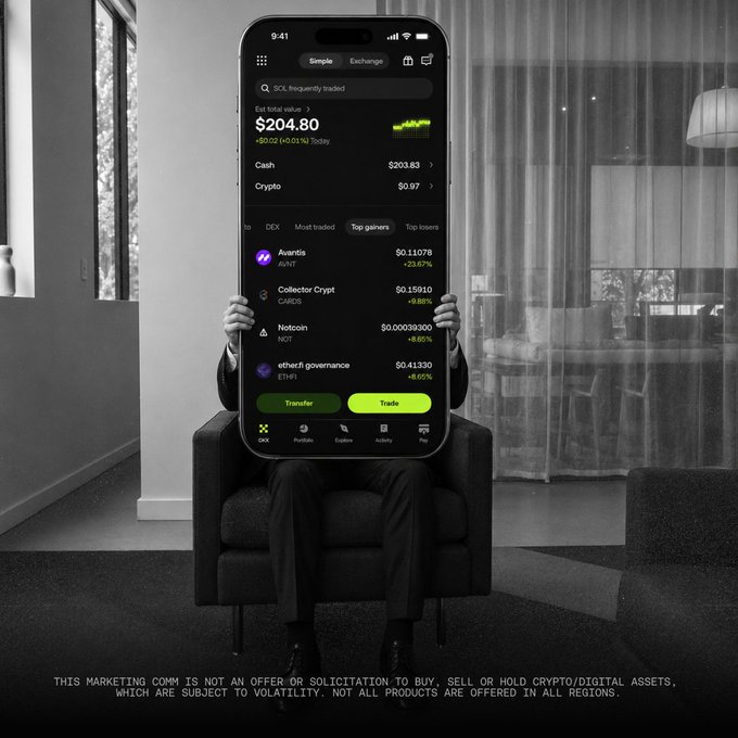A person seated in an armchair holds a large phone displaying the OKX exchange app, featuring a portfolio total of $204.80 and crypto holdings in AVANTIS, CARROS, NOY, and ETHFI. The image promotes downloading the OKX trading platform, which carries a disclaimer noting the marketing is not an offer to buy or sell crypto/digital assets subject to volatility.