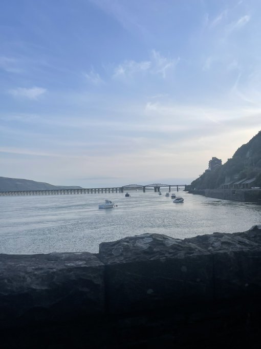 A serene waterfront view showing a long bridge stretching across the water with small boats, framed by hills and a stone wall in the foreground.