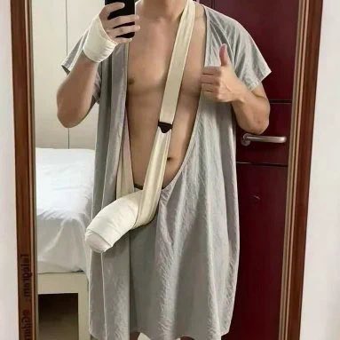 A mirror selfie of a person wearing a hospital gown with their arm in a sling and a bandage on their head, giving a thumbs-up.