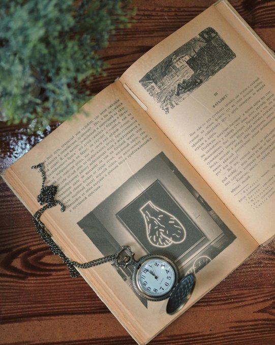 An open vintage book resting on a wooden surface with a pocket watch on a chain draped across its pages. The book appears to be in a Slavic language and contains illustrations.