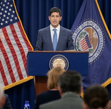 A man in a dark suit and blue tie stands at a podium bearing the Federal Reserve seal, speaking in front of an American flag and a Board of Governors flag.