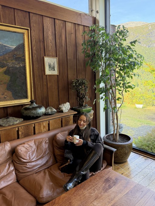 A woman in a brown sweater and black boots sitting on a leather couch holding a cup, with a large potted plant and wood-paneled walls in a rustic interior.