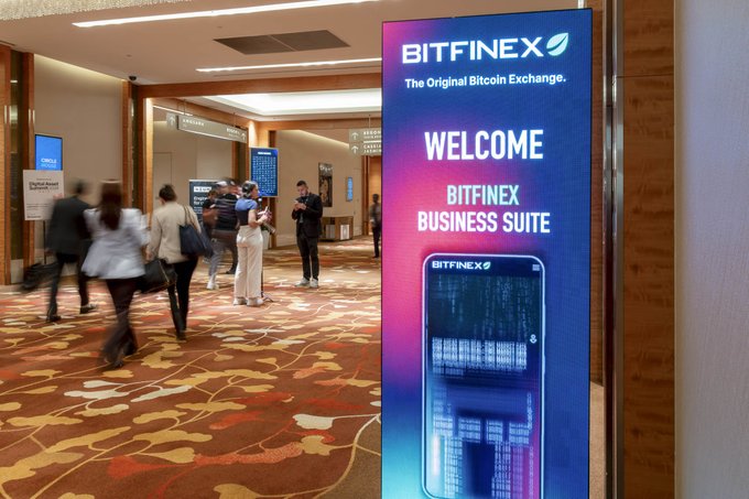 A large illuminated "BITFINEX" sign reading "The Original Bitcoin Exchange" and "WELCOME BITFINEX BUSINESS SUITE" displays prominently in a hotel hallway, where attendees are seen gathering outside the suite following the Token2049 Singapore conference. The scene captures Bitfinex's hospitality presence at the crypto event, providing a dedicated space for clients to discuss emerging topics like real-world assets and tokenized stocks mentioned in the accompanying post.