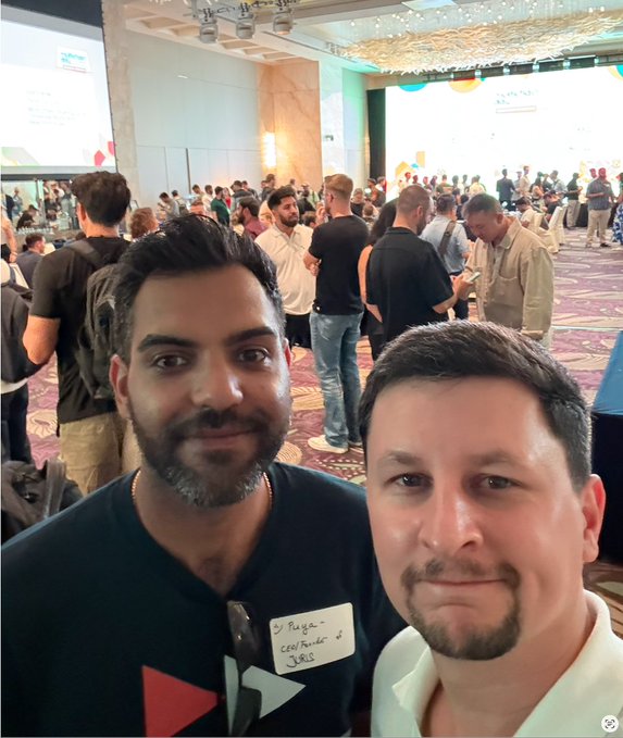 Selfie of two men at a crowded conference or meetup event; one wears a name tag reading "Jorge - CEO/Founder - JAR3S," and presentation screens are visible in the background.