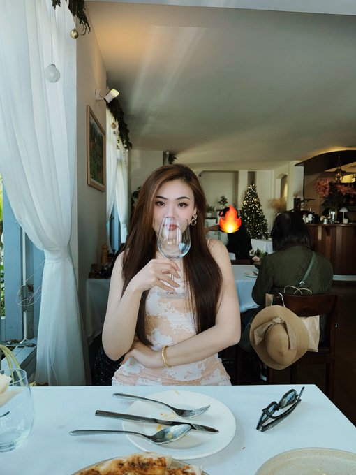 The image shows a woman sitting at a table with a plate of food in front of her. She has a wine glass in her hand, which she appears to be drinking from or about to drink. The background reveals that there are other people present as well. A Christmas tree can also be seen in the room, adding to the festive atmosphere.