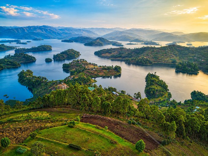 The image shows a beautiful view of a lake surrounded by mountains and lush green fields. The sky above the scene is blue with some clouds scattered throughout. There are several small islands located within the lake, adding to its picturesque charm. In the foreground, there's an aerial perspective that captures the vastness of the landscape, emphasizing the scale and grandeur of nature.