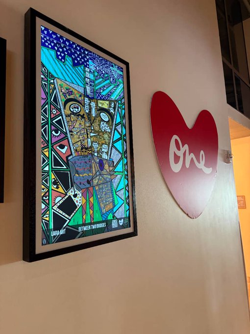 The image features a colorful painting of a face hanging on a wall, with a heart-shaped sign underneath that reads "ONE" in white letters. The painting depicts an abstract representation of a person's face, while the heart-shaped sign is a simple yet striking design. Both elements are displayed against a beige wall, creating a visually appealing contrast between the vibrant colors of the painting and the stark white of the sign.