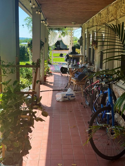 A sunlit outdoor porch with terracotta tile flooring, lined with bicycles and potted plants, leading toward a grassy yard with a white canopy.