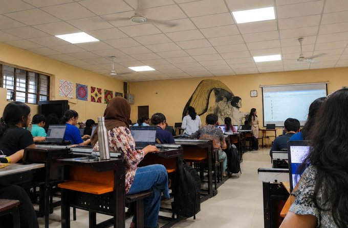 Students sit at desks with laptops in a classroom, facing a teacher standing near a whiteboard and a decorated bulletin board.