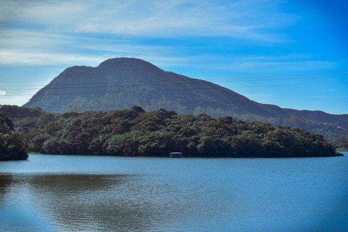 The image features a mountain with a lake at its base, surrounded by trees. In the foreground of the photo, there's an island that appears to be a small cabin or house. The sky above the scene has some clouds scattered across it, adding depth and atmosphere to the landscape.